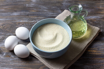 A blue ceramic bowl with semolina, three fresh white eggs and a glass bottle of vegetable oil on a rustic wooden table