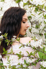 Naklejka premium Young brunette woman with long wavy hair wearing a white dress is posing near a blooming cherry tree in a spring garden, enjoying the scent of flowers and the warmth of the sun