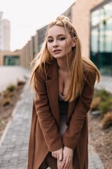 Influencer girl with braided blonde hair, wearing a stylish brown coat, leaning forward and posing...