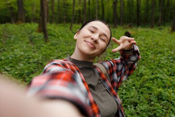 Young female hiker is taking a selfie while making a peace sign in a lush green forest, enjoying...