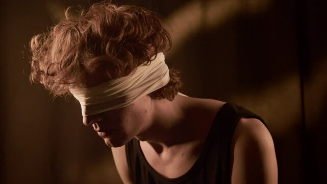 Chest-up shot of red-haired blindfolded young man dressed in black tank top slowly moving his head realising he cant see