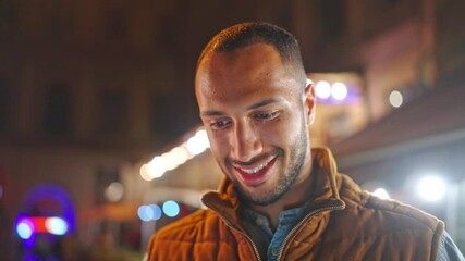Portrait of African American man looking down and positively smiling. Handsome bearded male considering something while standing outdoors. In blurred background shining lights. Night city concept.