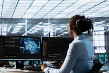 Engineer working at PC desk, doing checkup on AI server room providing computing resources. African american IT professional investigating data center mainframes using computer