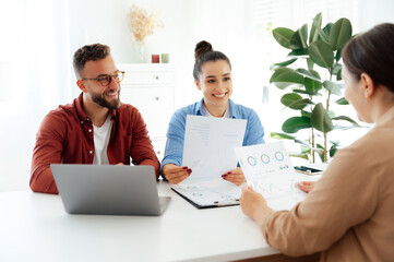 Two positive coworkers are holding papers with business charts, showing them to a third colleague,...