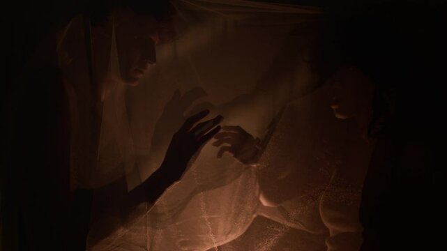 Young couple behind translucent veil reaching toward each other in delicate manner, warm diffused light casting gentle shadows of their hands