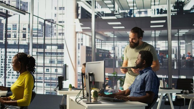 Manager coming to check on IT employee at office desk testing and deploying programs and systems. Team leader supervising developer writing code on computer in workspace, camera B
