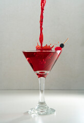 Red liquor stream splashing into cherry cocktail in conical glass on ceramic table against grey background