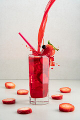 stream of red liquor splashing into strawberry cocktail with ice in crystal glass on ceramic table surrounded by strawberry slices against grey background