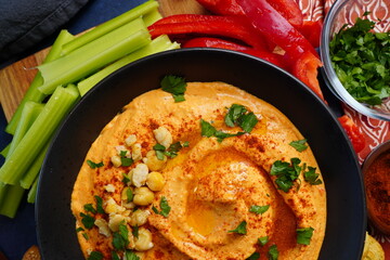 Making red pepper hummus with fresh ingredients and herbs.  Roasted Red Pepper hummus made with chickpeas, olive oil, parsley, paprika, garlic and tahini.  Sesame sticks, celery, bread and red peppers