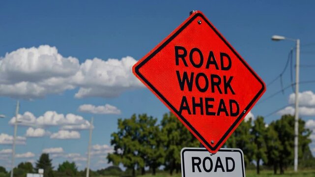 Road Work Ahead Sign on a Summer Afternoon: Safety Warning for Drivers on a Clear Highway Surrounded by Greenery Under a Bright Blue Sky