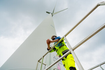 Worker inspects wind turbine at a renewable energy site during overcast skies, ensuring safety and...