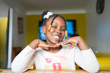 Una bambina africana allegra e sorridente posa in modo giocoso con le mani sotto il mento, esprimendo felicità e sicurezza. I suoi capelli sono acconciati con trecce decorate con perline dorate 
