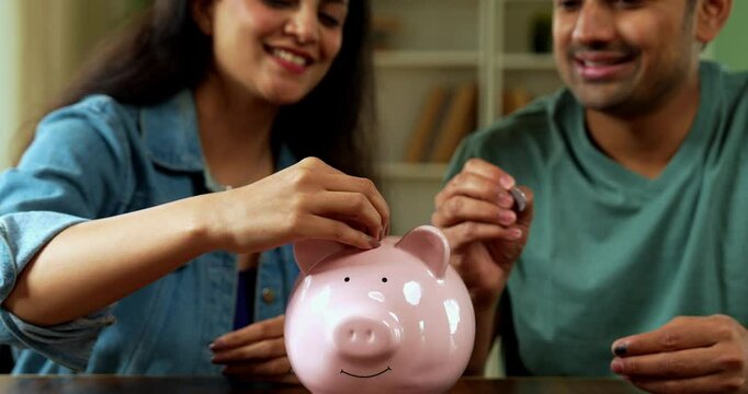 Indian Asian young couple in their twenties sitting at a home desk, happily saving money in a piggy bank, planning for their future, managing finances, and securing financial stability together.