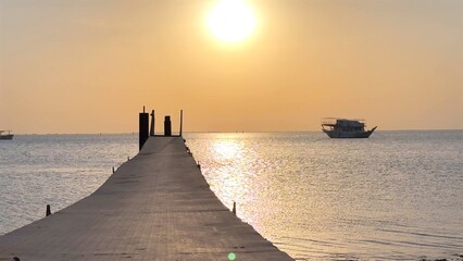 Rustic dock reaches into still ocean beneath warm sunset hues, anchored boat in distance adds nautical charm, soft reflections evoke sense of calm and timeless wanderlust.