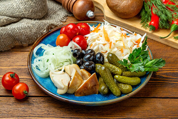 Assorted snacks: pickled cucumbers, marinated mushrooms, sauerkraut in a plate on a wooden background.