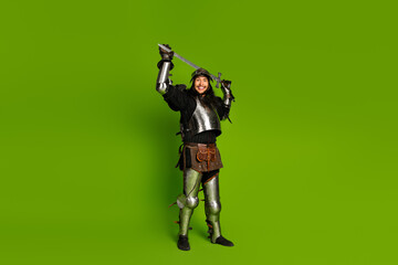 Young man in medieval knight armor posing with sword on green background.