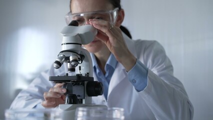 Woman scientist wearing lab coat and protective glasses is using microscope in modern laboratory...