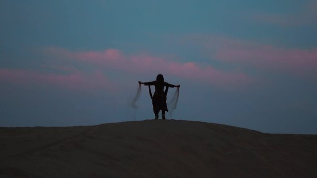 Silhouette of a young Indian girl playing with sand during sunset at Sam Sand Dunes in Thar desert at Jaisalmer, Rajasthan, India. Tourist enjoying vacation. Girl throwing sand in air.
