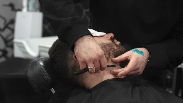 Professional barber carefully shaving client's beard with straight razor, applying gel and using hot towel for precise grooming in contemporary barbershop setting