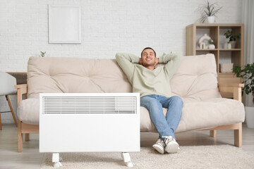 Fototapeta premium Young man sitting on sofa near electric heater in living room