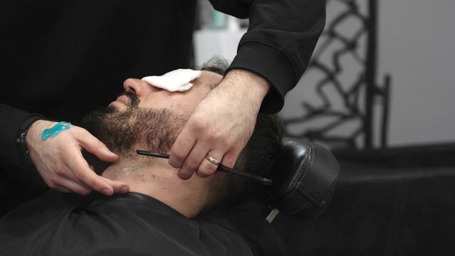 Professional barber carefully shaving client's beard with straight razor, applying gel and using hot towel for precise grooming in contemporary barbershop setting