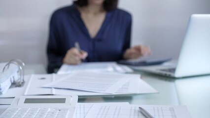 Magnifying glass, white calculator, pen and documents are lying on accountant's desk. Businesswoman wearing blue dress is working with documents on the background. Business and audit concept