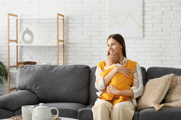 Young mother with little baby in sling sitting on sofa in living room