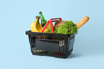 Black shopping basket full of different fresh products and baguette on blue background