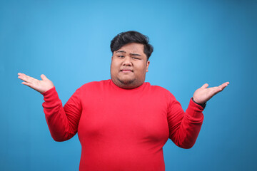 Fototapeta premium Portrait of Young Asian Fat Guy Showing Shrugging Hands Gesture Isolated on Blue Background
