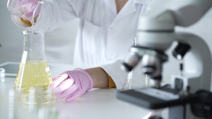 A scientist, wearing a lab coat and pink protective gloves, is pouring a yellow oily liquid from one beaker to another near microscope in laboratory, close up. Medicine and science