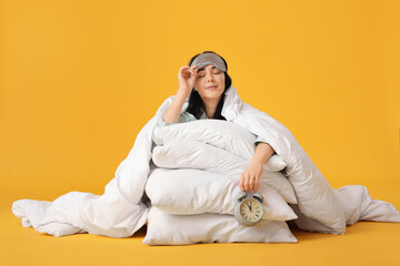 Pretty young woman with mask, pillows and alarm clock sleeping on yellow background