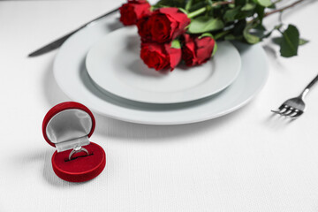 Engagement ring in box, flowers and tableware on white tablecloth, closeup. Table setting for...