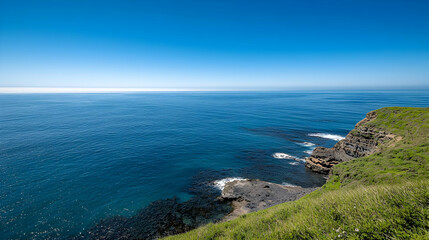 Obraz premium Vast Deep Blue Ocean Meets Rugged Cliff with Green Vegetation Under Clear Sky