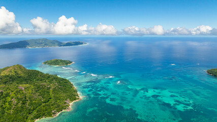 Lush green islands surrounded by clear turquoise ocean under a bright blue sky with white clouds. Seychelles, Mahe.