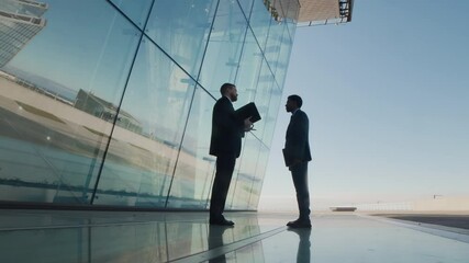 Low angle slow motion shot of two suited men, holding documents, discussing business outside modern glass building reflecting bright blue sky and surrounding cityscape
