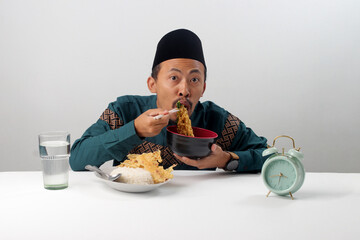 Asian Muslim man lifting instant noodles with chopsticks from a black bowl, smiling at the camera during Suhoor, the pre-dawn meal before fasting in Ramadan