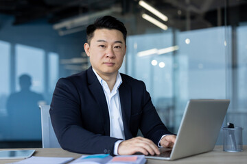 Portrait of a serious young Asian man, office worker, company owner, working in the office at a laptop and looking confidently at the camera