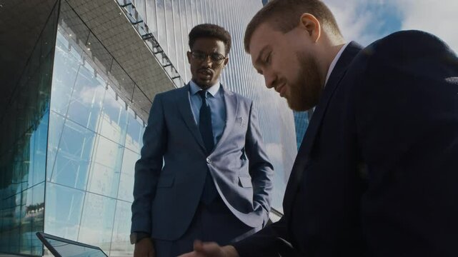 Low angle shot of African American man in formal two-piece suit helping his colleague with difficulties with laptop outside tall modern glass building in business district