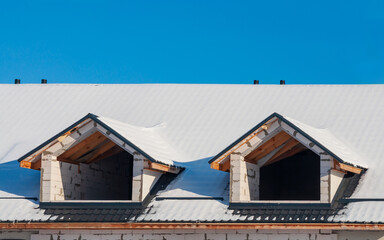 Snowy rooftop with dormer windows on an unfinished construction site against a bright blue sky.
