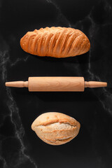 A freshly crunchy baked loaf of bread on a black marble surface with a rolling pin. Flat lay.