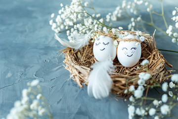 Two chicken eggs with hand drawn happy faces with wreaths in a nest of dry grass with feathers and...