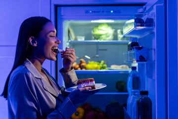 Hungry young woman in pajamas eating sweet cake late at night, standing near open refrigerator....