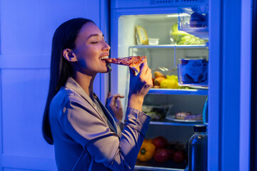 Hungry lady in sleepwear biting slice of pizza, standing near open refrigerator in kitchen late at night, enjoying fast food snack