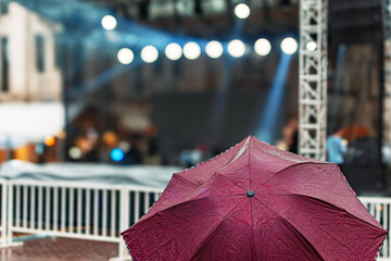 a person holding a maroon color umbrella in the rain against covered outdoor stage concert music...
