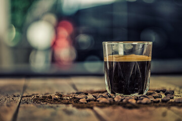 A warm-toned photo of espresso in a glass, surrounded by coffee beans and grounds. Dark and warm.
