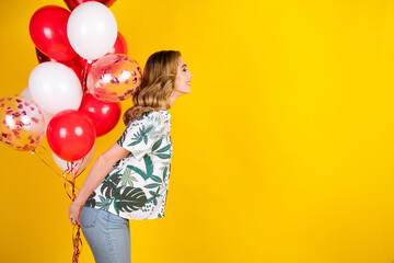 Stylish young woman holding festive balloons against a vibrant yellow background, embracing...