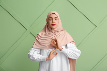 Beautiful young Muslim woman in hijab meditating on green background. Zen concept
