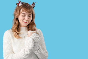 Pretty young woman in mittens holding cup of hot chocolate with marshmallows on blue background