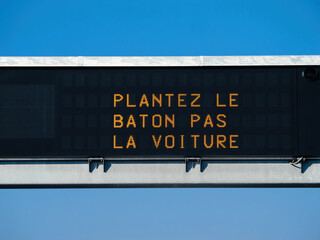 A digital highway sign in clear blue skies displays the messagePlantez le baton pas la voiture, encouraging environmental consciousness and alternative transport methods