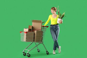 Young woman with houseplant and shopping cart full of cardboard boxes on green background